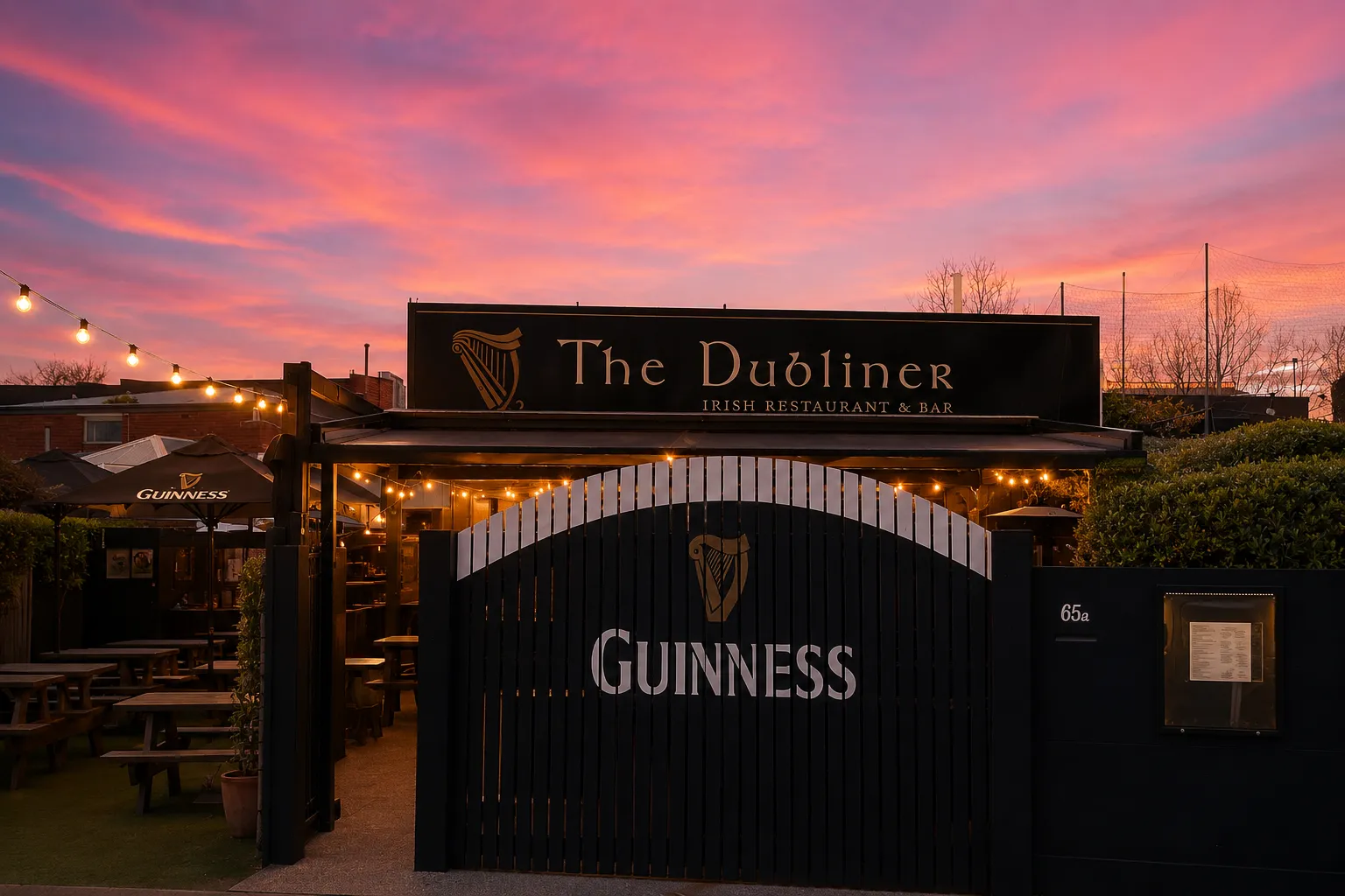 The Dubliner at dusk — the Celtic sign lit gold beneath a pink sunset, string lights and Guinness gates over the beer garden